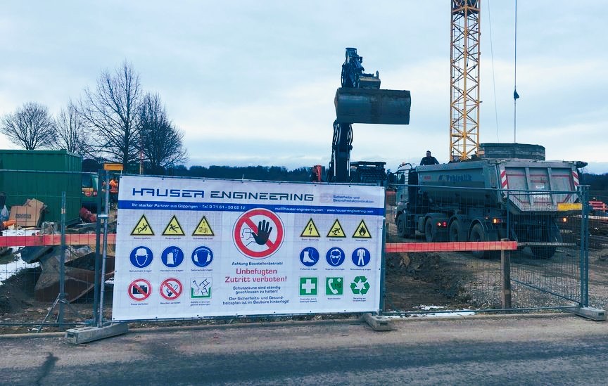 Bauschild von HAUSER ENGINEERING mit Sicherheits- und Gesundheitshinweisen am Bauzaun einer Baustelle